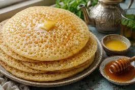 طريقة-عمل-خبز بغرير جزائري ذهبي مليء بالفقاعات الكثيفة، مكدس في طبق ومزين بالعسل السائل والزبدة الذائبة، مع إبريق شاي النعناع
