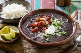 طبق شوربة فخاري كوبي يحمل شوربة الفاصوليا السوداء الداكنة الكثيفة، تعلوها مكعبات اللحم المقدد المدخن والبصل المفروم والكزبرة الخضراء، مع ملعقة من الكريمة الحامضة البيضاء وأرز أبيض جانبي وشرائح الليمون، على طاولة خشبية ملونة بأجواء هافانا القديمة