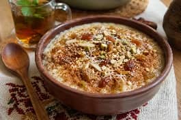 طبق تقديم فخاري مليء بأرز بالحليب المصري الكريمي، وجهه ذهبي مائل للبيج ومزين برشة من القرفة المطحونة والزبيب الذهبي المنتفخ وجوز الهند المبشور والمكسرات المحمصة، مع ملعقة خشبية على مفرش مصري تقليدي بجانب كوب شاي بالنعناع