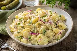 طبق سلطة بطاطا ألمانية باردة، قطع بطاطا طرية مغطاة بصوص المايونيز والخردل الكريمي، مع شرائح البصل الأحمر، مكعبات الخيار المخلل، البقدونس الطازج، ورشة من الفلفل الأسود، في طبق تقديم سيراميكي على طاولة حديقة