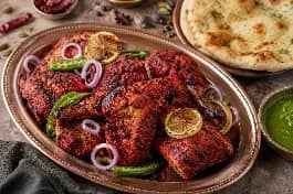 طبق نحاسي يحمل أفخاذ دجاج تندوري مشوية بلون أحمر قانٍ وبقع سوداء من الفرن، يعلوها شرائح ليمون مشوي وبصل أحمر وحلقات فلفل أخضر حار، بجانب صلصة نعناع خضراء وشرائح نان بالزبدة، على خلفية من التوابل الهندية المبعثرة