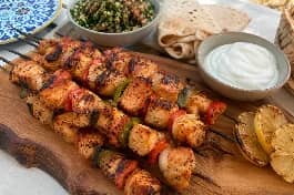 طبق تقديم خشبي يحمل أسياخ شيش طاووق الدجاج اللبناني المشوية على الفحم، قطع الدجاج الذهبية المتبلة بصوص الليمون والثوم تلمع وتتخللها قطع الفلفل الحلو والبصل، بجانب طبق من الثومية البيضاء الناعمة، خبز عربي محمص، وشرائح ليمون مشوي، على مائدة لبنانية صيفية بجانب سلطة تبولة