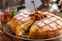 بسطيلة دجاج مغربية ذهبية كبيرة في صينية فرن، سطحها مرشوش بالسكر البودرة والقرفة المطحونة بنمط شبكي، مزينة باللوز المحمص والفواكه الجافة، بجانبها إبريق شاي مغربي وكؤوس زجاجية ملونة