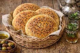 طريقة-عمل-أرغفة خبز الدار المغربي ذهبية اللون ومستديرة مع رشة سميد على الوجه وبذور اليانسون، موضوعة في سلة من الخيزران مع كؤوس شاي نعناع