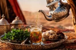 طقس تقديم شاي بالنعناع والزعفران المغربي، إبريق شاي فضي مزخرف يصب الشاي الذهبي من ارتفاع في كأس زجاجية صغيرة مزينة بورق النعناع وخيوط الزعفران، مع حلويات مغربية على مائدة صحراوية