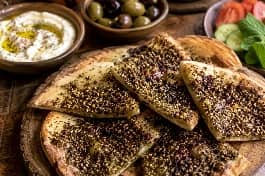 أرغفة خبز طابون فلسطيني محمصة ومغطاة بزيت الزيتون والزعتر الأخضر مع السماق الأحمر، تقدم على طبق خشبي تقليدي مع زيتون ولبنة، مشهد دافئ ومليء بالتراث