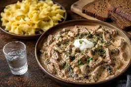 طبق تقديم عميق يحمل ستروغانوف اللحم الروسي الكلاسيكي، قطع لحم بقري طري في صوص كريمة بالفطر البني الذهبي، مزين بالبقدونس الطازج والقشدة الحامضة، بجانب طبق من المعكرونة البيضاوية العريضة، وطاولة خشبية داكنة بجانب كأس فودكا مثلجة وخبز الجاودار