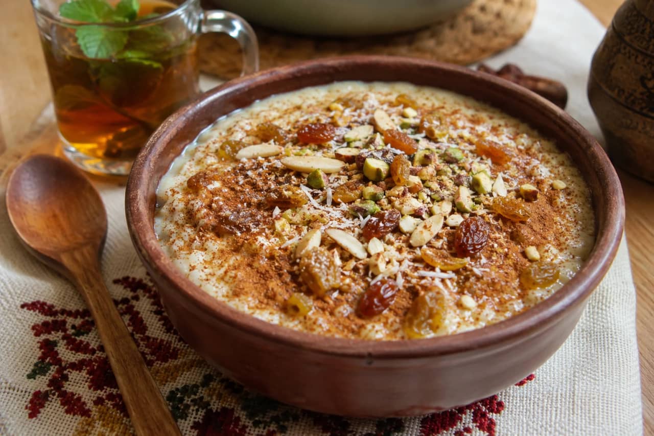 طبق تقديم فخاري مليء بأرز بالحليب المصري الكريمي، وجهه ذهبي مائل للبيج ومزين برشة من القرفة المطحونة والزبيب الذهبي المنتفخ وجوز الهند المبشور والمكسرات المحمصة، مع ملعقة خشبية على مفرش مصري تقليدي بجانب كوب شاي بالنعناع
