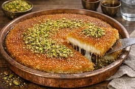 طريقة-عمل-طبق كنافة خشنة فلسطينية ذهبية مقرمشة مع طبقة قشطة وجبن نابلسي ذائب، مزينة بالفستق الحلبي والقطر، تقدم في صينية نحاسية