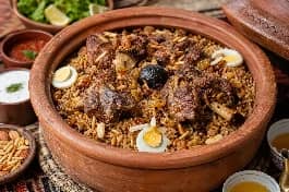 طبق رز حساوي سعودي في قدر فخاري، أرز بني مدخن مفلفل مع قطع لحم ضأن متبلة بالبهارات الخليجية واللومي، مزين بالمكسرات والزبيب على مائدة تراثية أحسائية