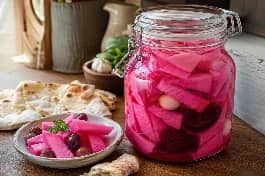 برطمان زجاجي كبير من مخلل اللفت الوردي الشامي المقطع، قطع لف مثلثة وشرائح بنجر حمراء وفصوص ثوم تطفو في محلول الخل الزهري، مع طبق جانبي من الزيتون والخبز العربي
