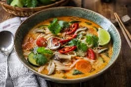 وعاء شوربة توم يام تايلاندية نباتية، مرق برتقالي كريمي مع خضار ملونة، فطر، نودلز أرز، وزيادة من حليب جوز الهند، مزينة بأوراق الكزبرة الطازجة، شرائح الفلفل الحار، وقطرات من زيت الفلفل الحار على طاولة خشبية