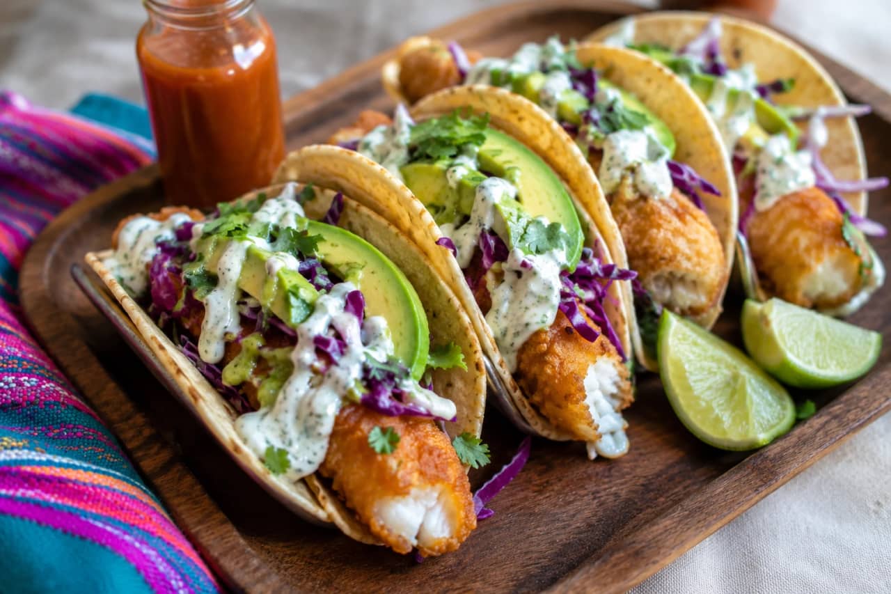 طبق خشبي يحمل سلالاً من تاكو السمك المكسيكي الذهبي المقرمش، محشوة بقطع فيليه سمك أبيض مقلية وشرائح أفوكادو كريمية وملفوف بنفسجي مقرمش، يعلوها صوص الكريمة البيضاء الحارة وخطوط من صوص الأفوكادو الأخضر، بجانب شرائح ليمون أخضر وصوص حار، على مفرش مكسيكي ملون