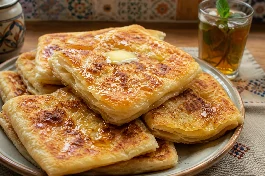 طريقة-عمل-خبز مسمن جزائري مورق وذهبي مربع الشكل، مدهون بالزبدة والعسل ومكدس في طبق تقديم مع كأس شاي النعناع