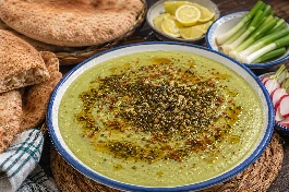 طريقة-عمل-طبق بصارة مصرية بقوام مخملي أخضر فاتح، مزينة بتقلية مدخنة من الثوم والكزبرة الجافة والشبت، مع رشة من الشطة الحمراء، ويحيط بها خبز بلدي أسمر وفصوص ليمون وشرائح بصل أخضر