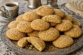 طريقة-عمل-أقراص كحك عجل بلدي مصرية ذهبية اللون ومستديرة، مزينة بحبات السمسم المحمص، مرصوصة في طبق كبير، بجانبها فنجان قهوة تركي، وبعض الأقراص مكسورة تظهر قوامها المبروم الهش
