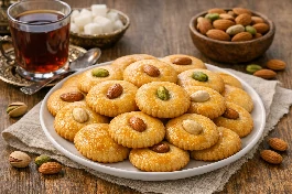 طريقة-عمل-قطع بسكويت سادة مصرية ذهبية اللون ومستديرة، مرصوصة في طبق تقديم، بجانبها فنجان شاي أحمر، بعض القطع مزينة بحبة لوز أو فستق في المنتصف