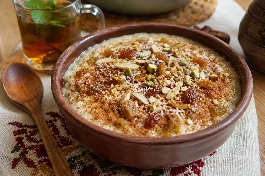 طبق تقديم فخاري مليء بأرز بالحليب المصري الكريمي، وجهه ذهبي مائل للبيج ومزين برشة من القرفة المطحونة والزبيب الذهبي المنتفخ وجوز الهند المبشور والمكسرات المحمصة، مع ملعقة خشبية على مفرش مصري تقليدي بجانب كوب شاي بالنعناع