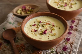 طبق فخاري صغير (شيكورا) يحمل أرز باللبن الهندي (فيرني) بحبيباته الناعمة، مبرد ومزين برقائق اللوز الذهبي والفستق الأخضر المفروم وخيوط الزعفران وبتلات الورد المجففة، مع ملعقة خشبية صغيرة على مفرش مطرز هندي
