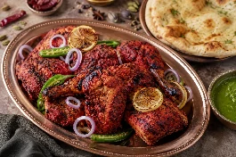 طبق نحاسي يحمل أفخاذ دجاج تندوري مشوية بلون أحمر قانٍ وبقع سوداء من الفرن، يعلوها شرائح ليمون مشوي وبصل أحمر وحلقات فلفل أخضر حار، بجانب صلصة نعناع خضراء وشرائح نان بالزبدة، على خلفية من التوابل الهندية المبعثرة