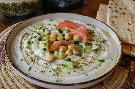 طبق سلطة طحينة كويتية بيضاء كريمية، مزينة بحبات الحمص الذهبية، مكعبات الخيار الطازج، شرائح الطماطم، رشة من البقدونس والكمون، مع خبز عربي محمص على مائدة كويتية تقليدية