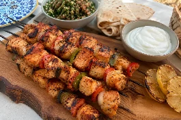 طبق تقديم خشبي يحمل أسياخ شيش طاووق الدجاج اللبناني المشوية على الفحم، قطع الدجاج الذهبية المتبلة بصوص الليمون والثوم تلمع وتتخللها قطع الفلفل الحلو والبصل، بجانب طبق من الثومية البيضاء الناعمة، خبز عربي محمص، وشرائح ليمون مشوي، على مائدة لبنانية صيفية بجانب سلطة تبولة