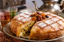 بسطيلة دجاج مغربية ذهبية كبيرة في صينية فرن، سطحها مرشوش بالسكر البودرة والقرفة المطحونة بنمط شبكي، مزينة باللوز المحمص والفواكه الجافة، بجانبها إبريق شاي مغربي وكؤوس زجاجية ملونة