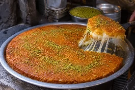 صينية كنافة نابلسية خشنة فلسطينية ذهبية اللون، مقلوبة ليظهر الوجه المقرمش باللون البرتقالي الذهبي، يعلوها الجبن الأبيض الذائب والمطاط، ومزينة بالفستق الحلبي الأخضر المطحون والقطر اللامع، على صينية معدنية كبيرة في ورشة كنافة تقليدية في نابلس
