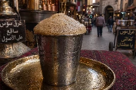 كوب معدني تقليدي لمشروب عرق السوس البارد مع رغوة كثيفة على الوجه، بخلفية تراثية لسوق شعبي في رمضان