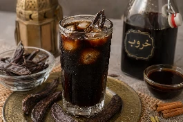 كوب زجاجي طويل من مشروب الخروب البارد الداكن مع مكعبات ثلج وحبات خروب كاملة للتزيين على طاولة رمضانية