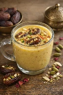 طريقة-عمل-كوب زجاجي شفاف يحتوي مشروب حليب ذهبي دافئ مزين بخيوط الزعفران، مع تمرات محشوة بالمكسرات وحبات فستق ولوز تطفو على الوجه، على خلفية دافئة
