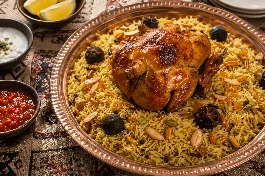 طبق تقديم نحاسي كبير يحمل كبسة دجاج سعودية ذهبية اللون، يتصدره دجاجة كاملة مشوية بالفرن بلون محمر لامع، الأرز البسمتي طويل الحبة مفلفل ومتبل بالزعفران والهيل والقرنفل، مزين باللوز المحمص والزبيب الذهبي وشرائح الليمون المجفف (لومي)، على مفرش عربي تقليدي بجانب طبق سلطة الدقوس الحارة