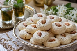 أقراص غريبة تونسية باللوز وماء الزهر بلون أبيض ثلجي مزينة باللوز الكامل، تقدم في طبق تقديم فاخر مع كؤوس شاي أخضر بالنعناع وزهور بيضاء